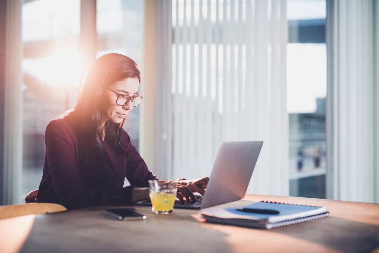 En kvinne sitter ved et skrivebord på et kontor og jobber på en bærbar PC med notatbok og mobil ved siden av seg. Bildet illustrerer en profesjonell arbeidssituasjon som passer i kontekst med innhold om hva er revisjon og kontroll av økonomiske prosesser.